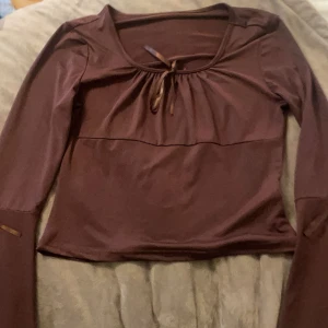 Brun långärmad topp med knyt - Säljer en brun långärmad topp med utsvänga armar och snygg knytning vid halsringningen och armarna. Toppen har en mjuk, rundad form och är figurnära med en lätt rynkning framtill. Perfekt för dig som gillar enkla men stilrena detaljer. Pris kan diskuteras och köparen står för frakten