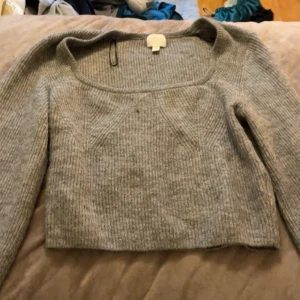 Grå ribbad stickad tröja - Säljer en grå stickad tröja med ribbad struktur och bred, fyrkantig halsringning. Tröjan är croppad och har långa ärmar, perfekt för dig som gillar en enkel men snygg look. Passar dig som vill ha något mjukt och bekvämt till hösten eller vintern. Pris kan diskuteras och köparen står för frakt