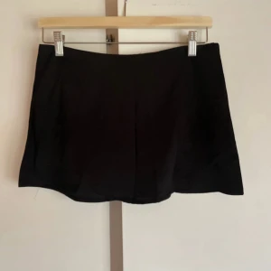 Svart kortkjol i bomull - En stilren svart kortkjol med enkel design och rak passform. Kjolen är tillverkad i mjukt bomullstyg och har en klassisk look utan synliga detaljer eller mönster. Perfekt för dig som gillar minimalistisk stil och vill ha en basic kjol i garderoben. Den har en dragkedja på sidan av höften!