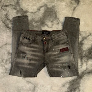 Grå Philipp Plein jeans med detaljer - Säljer ett par grå jeans från Philipp Plein med coola handgjorda detaljer som färgstänk i vitt och turkos, röda läderpatches med metallskalle och logga, samt unika nitar. Slim fit-modell med snygga slitningar och kontrastsömmar.