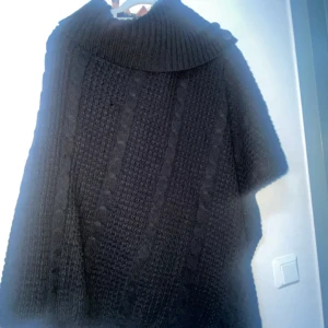 Svart stickad polotröja med kabelmönster - Svart stickad polotröja med bred ribbad krage och snygga kabelstickade detaljer framtill. Tröjan har en loose passform och är perfekt för kyliga dagar. Materialet är mjukt och tjockt, vilket gör den extra mysig och varm.