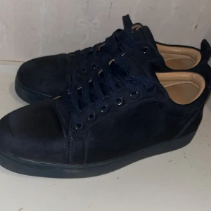 Svarta mockasneakers med röd sula - Snygga Mörkblåa  sneakers i mocka från Christian Louboutin med klassisk röd sula och mörkblåa snören. Skorna har rund tå och platt sula, samt beige innersida för extra kontrast. Perfekta för dig som vill sticka ut med en lyxig touch. Använt ett få tal Gånger, Pris kan diskuteras 
