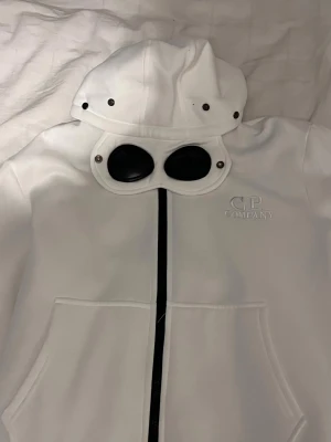 Vit goggletröja från C.P. Company - Säljer en vit gogglejacka från C.P. Company med ikoniska svarta glasögon på huvan och broderad logga på bröstet.