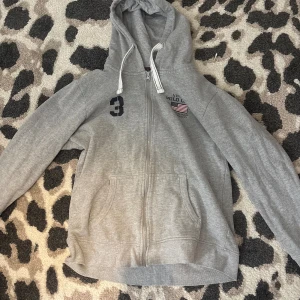 Grå hoodie från Vinson Polo Club - Grå hoodie med dragkedja från Vinson Polo Club, har huva med snörning och två fickor framtill. Broderad logga och siffra på bröstet. Insidan är mjuk och fleecefodrad, perfekt för kyliga dagar. Snygga detaljer med röd och svart rand på insidan av kragen.