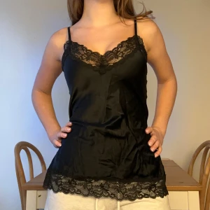 Svart Intimissimi sidentopp  - Säljer en svart linnetopp med tunna axelband och snygga spetsdetaljer vid halsringning och nederkant. Toppen har en mjuk, glansig yta och sitter skönt mot kroppen. Perfekt för dig som gillar en stilren look med lite extra edge. Använd 2 ggr och 100% siden därav priset💕 nypris: 600kr
