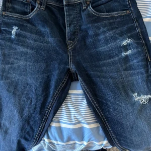 Mörkblå jeans med slitningar - Säljer ett par mörkblå jeans med snygga slitningar och tydliga kontrastsömmar. Klassisk femficksmodell med läderpatch bak i midjan. Jeansen har rak passform och är tillverkade i robust denim. Perfekta för en avslappnad och trendig look.