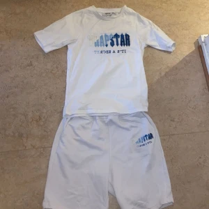 Vita Trapstar shorts med tryck - Säljer ett par vita shorts från Trapstar med blått och vitt tryck på ena benet. De har en avslappnad passform och resår i midjan för extra komfort. Perfekta för sommardagar eller chill hemma. Matcha med din favorit-tisha för en clean look.