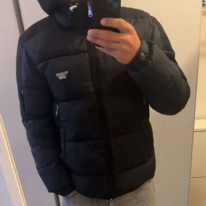 Svart pufferjacka alesandro zavetti - Svart pufferjacka från alesndro zavetti med huva och dragkedja framtill. Jackan har två sidofickor med dragkedja och en diskret zavetti-logga på bröstet. Perfekt för kalla dagar och har en modern, sportig look. Original pris 1299kr jag har storlek M och den passar jätte bra även när det är storlek L på den