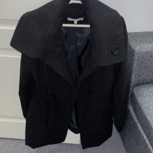 Svart trenchcoat med bred krage - Svart trenchcoat från Marks & Spencer med bred krage och stora knappar. Jackan har ett snyggt bälte i midjan, två fickor framtill och är helfodrad. Passformen är normal och modellen är långärmad, perfekt för kyliga dagar. Kappan nerifrån ser ut som en kjol vilket passar ifall man vill lyxiga till vardagen. Jackan är i storlek S/36