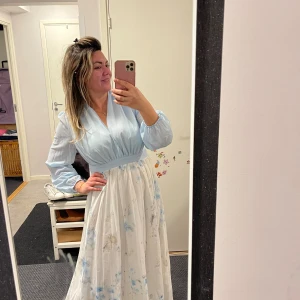 Ljusblå långklänning med blommigt mönster - Superfin långklänning med ljusblå överdel och v-ringning, samt långa ärmar med lätt puff. Kjolen är vit med diskret blommigt mönster i blått och beige. Klänningen har markerad midja och ett flowigt, romantiskt fall. Perfekt för dig som gillar en dreamy och feminin stil.Helt ny