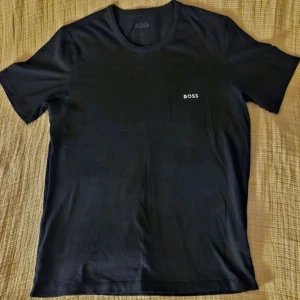 Svart t-shirt från BOSS, storlek M - Svart t-shirt från BOSS med stilren logotyp på bröstet. Klassisk rund hals och korta ärmar. Tillverkad i mjuk bomull som känns skön mot huden. Perfekt för dig som gillar enkel och snygg design med premiumkänsla.