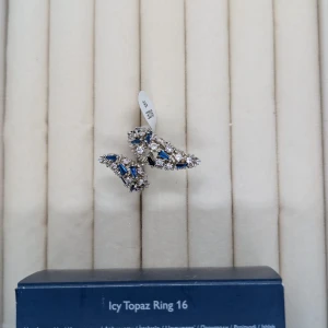 Icy Topaz silverring med blå stenar - Snygg silverring från Icy Topaz i storlek 16, dekorerad med glittrande vita och blå stenar i olika former. Ringen har en öppen design och kommer med en mörkblå satinpåse med silverdetaljer. Perfekt för dig som gillar bling och vill sticka ut.