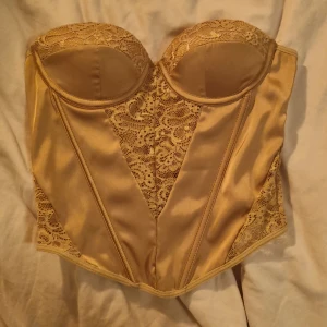 Guld korsett med spetsdetaljer - Snygg guld korsett med vadderade kupor och vackra spetsinslag framtill och på sidorna. Toppen har en smickrande form, dragkedja i sidan och är axelbandslös. Perfekt för att skapa en statement-look.