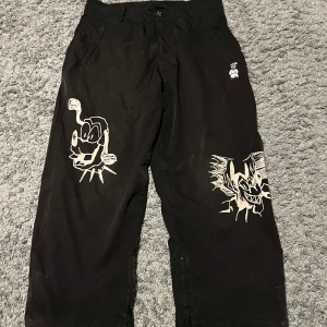 Svarta Harlaut täckbyxor - Unika svarta täckbyxor från Harlaut Apparel med vita grafiska tryck på benen och bakfickan. Byxorna har en loose passform, hög midja och är tillverkade i slitstarkt syntetmaterial. Perfekta för dig som vill sticka ut med coola detaljer och se steezy ut i backen. Pris kan diskuteras