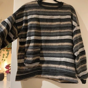 Retro randig tröja i svart, beige, vit och grå - Säljer en oversized tröja med breda ränder i svart, grå, vit och beige. Tröjan har långärmad passform och en stor ficka framtill. Mönstret är varierat med olika stickade strukturer som ger en cool vibe.