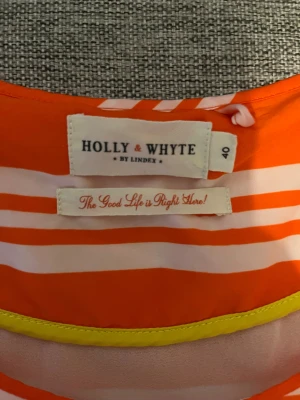 Randig orange klänning Holly & Whyte - Snygg ärmlös kortklänning från Holly & Whyte by Lindex i orange och vitt med randigt mönster. Klänningen har rund hals och en loose passform, perfekt för sommarens varma dagar. Materialet känns lätt och luftigt mot huden.
