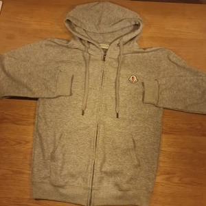 Grå Moncler hoodie med dragkedja - Snygg grå hoodie från Moncler med huva och snörning, hel dragkedja framtill och två fickor. Ikonisk Moncler-logga på bröstet. Mjuk bomullskänsla och ribbade muddar vid ärmslut och nederkant. Perfekt för chill eller streetwear.