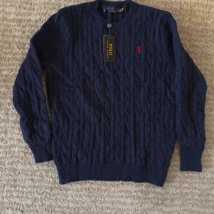 Marinblå stickad tröja Polo Ralph Lauren - Snygg marinblå stickad tröja från Polo Ralph Lauren med klassiskt kabelmönster och röd broderad logga på bröstet. Rund halsringning och ribbade muddar vid ärmslut och nederkant. Perfekt för dig som gillar stilren och tidlös design.m passar s.