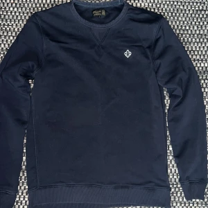 Mörkblå sweatshirt från Morris - Snygg mörkblå sweatshirt från Morris med rund hals och broderad logga på bröstet. Tröjan har ribbade muddar vid ärmslut och nederkant samt klassisk passform. Perfekt för dig som gillar stilrena och enkla plagg med diskreta detaljer.