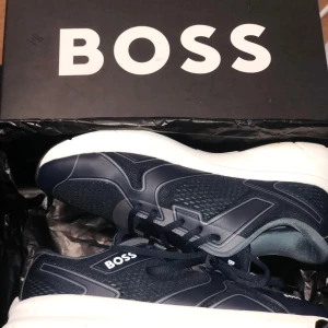 Marinblå sneakers från BOSS - Snygga marinblå sneakers från BOSS med vit platt sula och meshdetaljer. Skorna har snörning och rund tå, med BOSS-logga på sidan och tungan. Materialmix av syntet och textil ger en sportig vibe och skön komfort. Perfekta för dig som gillar stilrena och moderna sneakers. Nypris 2899kr mitt pris 1100kr hel nya oanvända!! Hugo boss skor är lite stora i st skulle kunna nästan passa nån med 41/5 eller kanske 42 