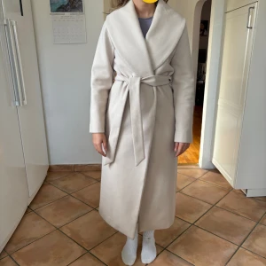 Beige  kappa med bälte - Snygg lång beige kappa med bred krage och omlottmodell. Kappan har ett matchande bälte i midjan, diskreta sidfickor och knappar på insidan. Materialet är mjukt och känns lyxigt, perfekt för dig som gillar stilrena och minimalistiska plagg. Extremt sparasqmt använd inga defekter och inte nopprig överhuvudtaget. Nypris 1099kr.