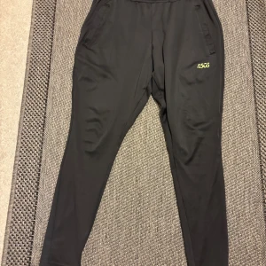 Svarta träningsbyxor 4505 i S - Svarta träningsbyxor från 4505 med smal passform och elastisk midja. Byxorna har diskreta fickor på sidorna och en gul 4505-logga på låret. Perfekta för gymmet eller chill hemma. Materialet är mjukt och stretchigt för maximal rörelsefrihet.