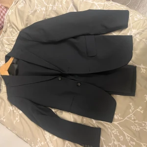 Svart kostym Lindbergh White slim fit - Säljer en svart kostym från Lindbergh White i slim fit-modell. Kavajen har två knappar framtill, klassiska slag och fickor med lock. Byxorna är raka och matchar kavajen perfekt. Kostymen är stilren och passar dig som gillar en smalare passform.