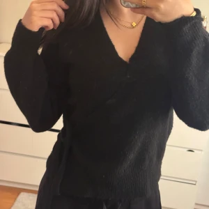 Svart omlott kofta med knyt - Mysig svart omlott kofta med knyt i sidan och v-ringad hals. Tröjan har långärmade ärmar och är stickad i ett mjukt material som känns skönt mot huden. 