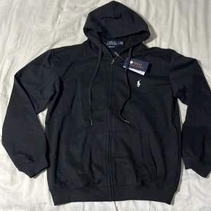Svart hoodie från Polo Ralph Lauren - Replika!!! Helt ny Passar storlek S Svart hoodie från Polo Ralph Lauren med dragkedja, huva med snörning och klassisk vit broderad logga på bröstet. Tröjan har ribbade muddar och två fickor framtill. Perfekt för en chill och stilren look.