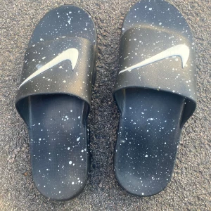 Svarta Nike sandaler med stänkmönster - Svarta sandaler från Nike med vitt stänkmönster och stor vit swoosh-logga på ovansidan. Sulan har en unik, upphöjd struktur för extra komfort och cool look. Tillverkade i syntetmaterial, perfekta för chill dagar och enkel att dra på.