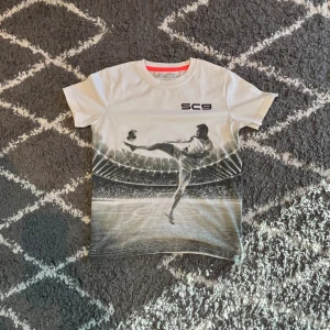 Vit t-shirt med fotbollstryck från SOC - Vit t-shirt från SOC med coolt fotbollstryck framtill där en spelare gör en kick på en stadion. SC9-logga på bröstet och kontrastfärgad röd insida vid halsen. Mjuk bomullskvalitet och klassisk passform med korta ärmar.