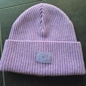 Ljuslila ribbstickad mössa Canada Snow - Säljer en ribbstickad mössa från Canada Snow i en snygg ljuslila färg. Mössan har uppvikt kant och en patch framtill med logga. Perfekt för kalla dagar och ger en trendig look till din outfit.