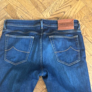 Jacob Cohën Bard blå jeans storlek 34 - Snygga blå jeans från Jacob Cohën, modell Bard. Klassisk femficksdesign med kontrastsömmar och knappgylf. Märkespatch i brunt läder baktill och unika detaljer på insidan. Passar dig som gillar stilrena och exklusiva jeans.