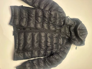 Svart dunjacka Peak Performance - Svart dunjacka från Peak Performance med huva och broderad logga på bröstet och huvan. Jackan har quiltad design, dragkedja framtill och sidofickor med dragkedja. Perfekt för kalla dagar och har en clean, sportig look. Kostar 2000 ny