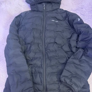 Svart Peak Performance pufferjacka - Svart pufferjacka från Peak Performance med huva och quiltad design. Jackan har dragkedja framtill och logga på bröstet samt ärmen. Perfekt för kalla dagar och har en clean, sportig look som funkar till det mesta.