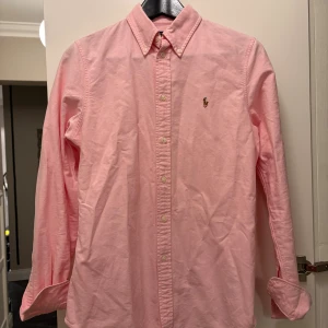Rosa skjorta från Polo Ralph Lauren - Snygg rosa skjorta från Polo Ralph Lauren i custom fit. Klassisk button-down krage, broderad logga på bröstet och knappar hela vägen. Tillverkad i mjuk bomull och har lång ärm. Perfekt för dig som gillar stilren och fräsch look.