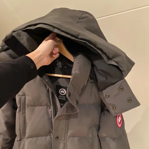 Grå dunjacka från Canada Goose - Säljer en grå dunjacka från Canada Goose med klassisk patch på ärmen. Jackan har stor huva, quiltad design och praktiska knappar samt dragkedja framtill. Perfekt för kalla dagar och har en clean, stilren look som funkar till allt. PRIS KAN DISKUTERAS, även kan du få fler bilder med den på eller liknande.