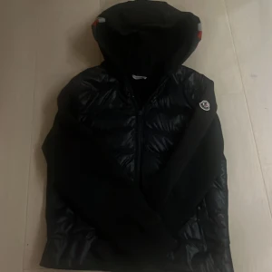 Mörkblå Moncler cardigan  - Mörkblå cardigan från Moncler med glansig pufferfront och mjuka ärmar. Jackan har huva, dragkedja och Moncler-logga på ärmen.  Nypris 7999kr vårt pris 2199. Den är i storlek 152 men skulle säga att den är lite liten i storleken.