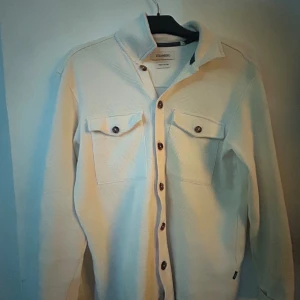 Vit overshirt från Strömberg - Snygg vit overshirt från Strömberg i bomull med grov struktur. Skjortan har klassisk krage, bruna knappar hela vägen och två stora bröstfickor med lock. Perfekt att slänga över en t-shirt för en chill look.