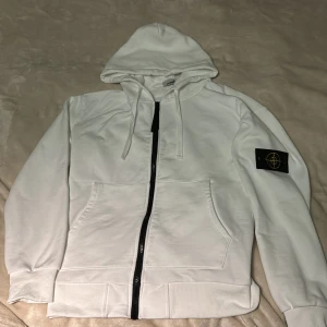 Vit hoodie från Stone Island - Vit hoodie från Stone Island med klassisk svart och gul patch på vänster ärm. Tröjan har huva med snörning, hel dragkedja framtill och två stora fickor. Tillverkad i mjuk bomull, perfekt för chill dagar. Snygg och stilren design med ribbade muddar.