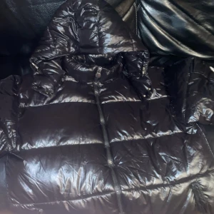Svart Moncler pufferjacka med huva - Säljer en svart pufferjacka från Moncler med glansig finish och huva. Jackan har dragkedja framtill, Moncler-logga på ärmen och praktiska fickor. Perfekt för kalla dagar och har en modern, oversized look som är riktigt snygg.