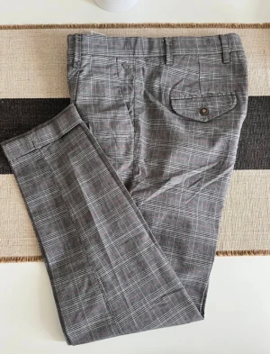 Rutig kostymbyxa Jack & Jones 30/32 - Snygga gråa kostymbyxor från Jack & Jones i rutigt mönster med diskreta röda linjer. Byxorna har carrot fit, bälteshällor, fickor med lock och knapp bak samt klassisk dragkedja. Perfekta för en stilren look med modern touch.