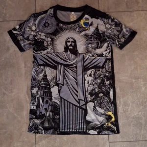 Svart och grå Brasilien fottbolls tröja  - Unik t-shirt med stort konstnärligt tryck av Kristusstatyn på framsidan, omgiven av detaljerade mönster och motiv i svart, grått och inslag av gult. Korta ärmar med tryck, svart baksida och rund hals. Perfekt för dig som gillar statement-plagg och coola prints.