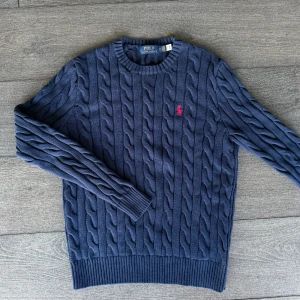 Marinblå Ralph lauren tröja  - Kabelstickad marinblå tröja från Polo Ralph Lauren med rund halsringning och klassisk röd broderad logga på bröstet. Tröjan är långärmad och har ribbade muddar vid ärmslut och nederkant. Perfekt för dig som gillar preppy och stilren look.