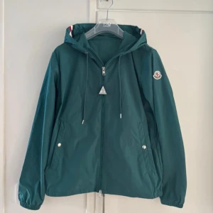 Grön vindjacka från Moncler med huva - Snygg grön vindjacka från Moncler med huva och coola rödvita detaljer på kragen. Jackan har dragkedja framtill, snörning vid huvan och två fickor med knappar. Perfekt för dig som gillar stilren design och vill ha något från ett exklusivt märke. Storlek 2 dvs s/m.