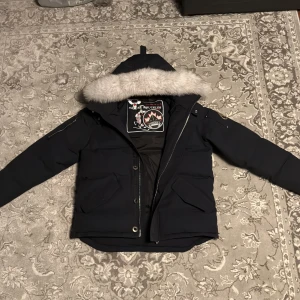 Moose Knuckles blå dunjacka med päls - Svart dunjacka från Moose Knuckles med stor fluffig ljusgrå päls på huvan, broderad hockeypatch på insidan och metallogo på ärmen. Jackan har dragkedja, knappar och två stora fickor framtill. Perfekt för kalla vinterdagar och riktigt snygg streetstil.