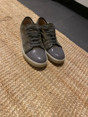 Grå sneakers från Lanvin med lackad tå - Snygga sneakers från Lanvin i mörkgrå mocka med grå lackad rund tå och vita sulor. Skorna har svarta snören och en stilren design som passar till jeans eller chinos. Materialmixen mellan mocka och lack ger en cool kontrast och exklusiv känsla. Skriv om du har några funderingar eller vill ha mer bilder. Har inte låda kanske Dustbag.