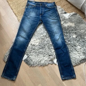 Jack and jones  jeans blå Grisch - Jack and jones jeans i blå tvätt med slim fit passform. Klassisk femficksdesign, guldfärgade nitar och tydliga slitningar framtill. Jeansen har normal midja och är gjorda i stretchigt denim för skön komfort. Perfekt för dig som gillar en smal siluett. Den har perfekta slitningar
