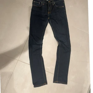Mörkblå jeans Nudie Skinny - Snygga mörkblå jeans från Nudie med skinny passform och klassiska orangea sömmar. Modellen har fem fickor, smal siluett och är tillverkad i stretchigt denim för extra komfort. Perfekt för dig som gillar en stilren och modern look.