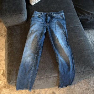 Blå jeans från Jack & Jones, 28/30 - Snygga blå jeans från Jack & Jones med klassisk femficksdesign och läderpatch bak i midjan. Jeansen har normal passform och raka ben, med coola slitningar framtill för en avslappnad vibe. Perfekt för dig som gillar stilrena och tidlösa jeans.